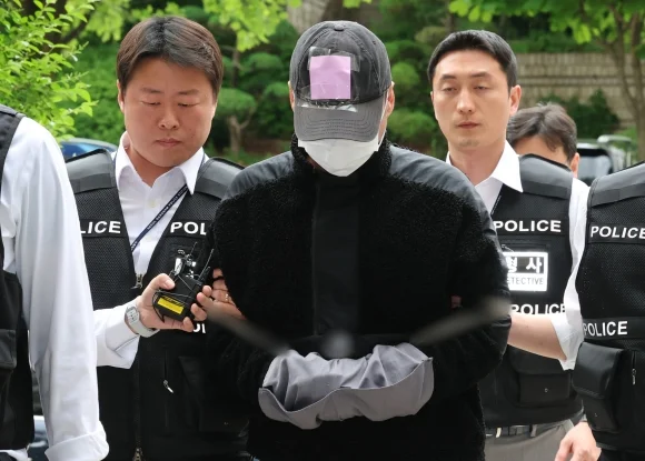 A person wearing a cap and mask is escorted by police officers outside a courthouse, with their hands bound and facing downward.
