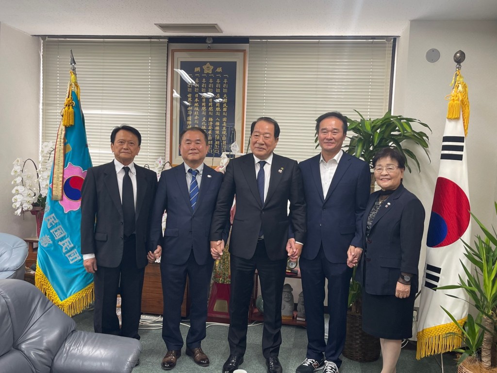 A group photo of Shinjuku Korean Association members, including chairman Jeong Jae-wook, posing together in an office with Korean flags and a potted plant.