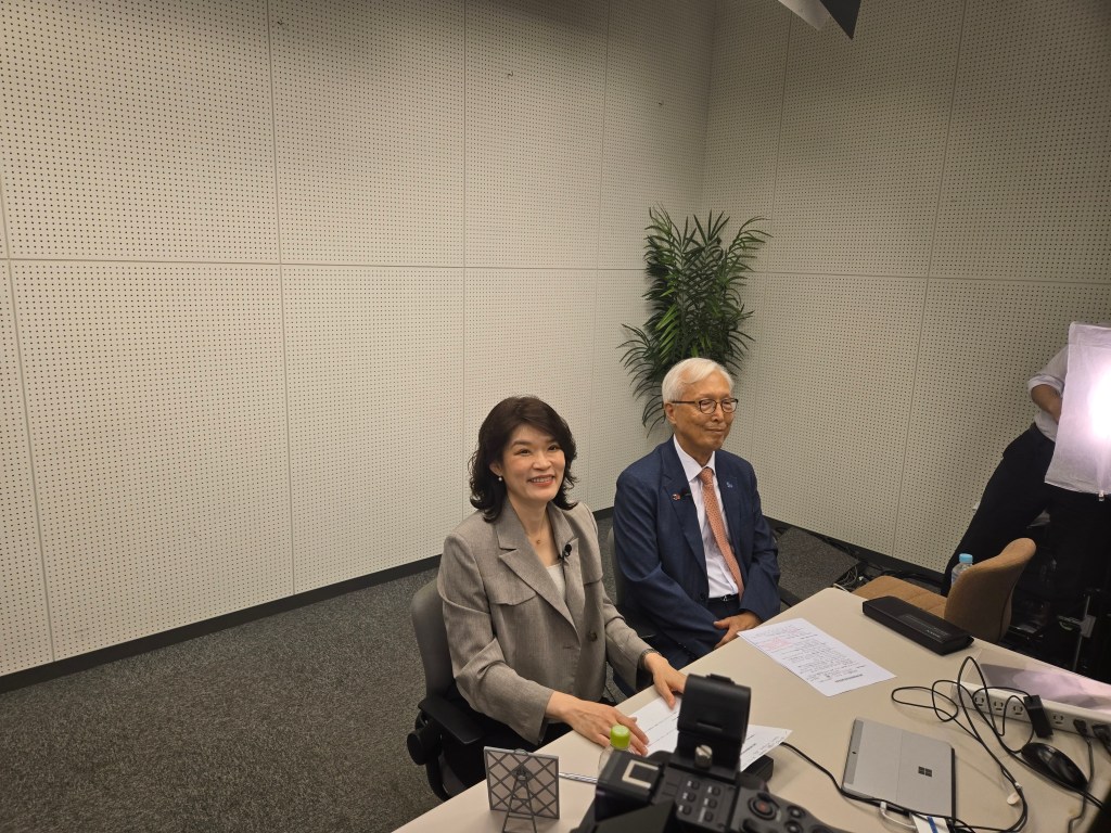 강연회에서 두 사람의 발표자가 앉아 있는 모습, 테이블 위에는 노트북과 문서가 놓여 있으며, 배경에는 식물이 보임.