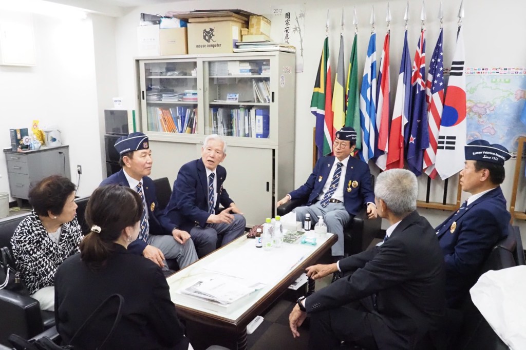 A meeting of Korean veterans in a conference room, with multiple national flags displayed in the background.