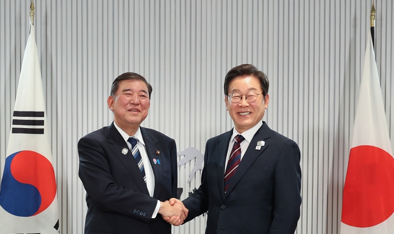 한국과 일본의 관계 강화를 위해 악수를 나누고 있는 두 남성, 한국과 일본 국기가 배경에 있다.