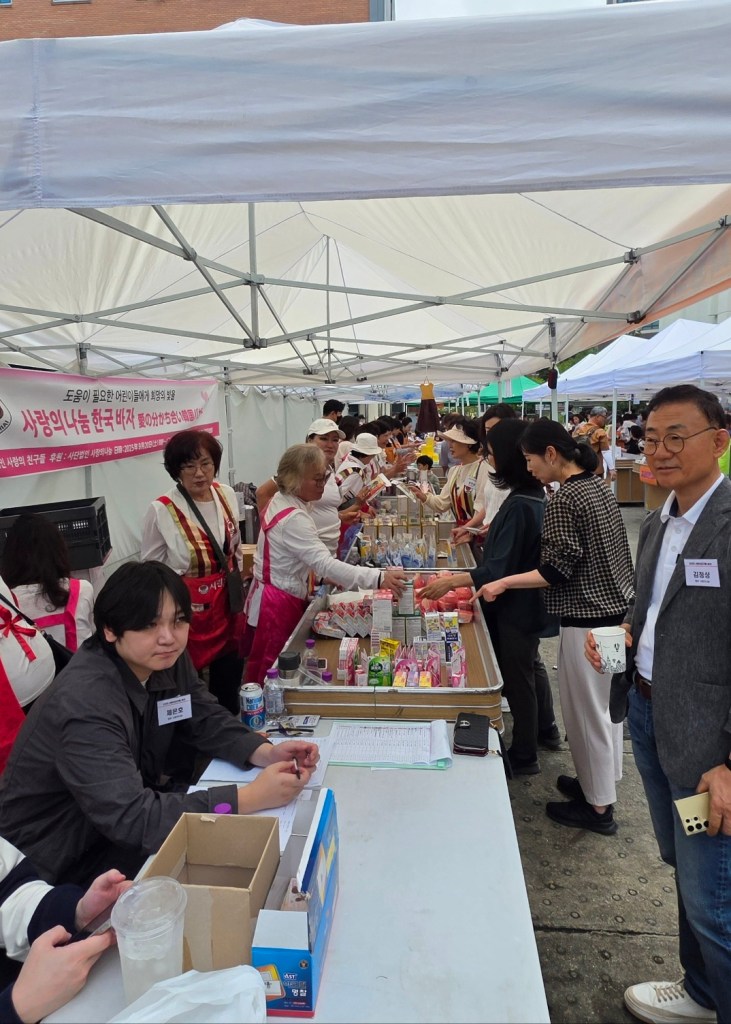 A group of volunteers is seen working at a booth during the 'Love Sharing Korea Bazaar' event, which aims to provide hope to children in need. Various traditional Korean foods and products are displayed on tables, with participants engaged in purchasing and interacting.