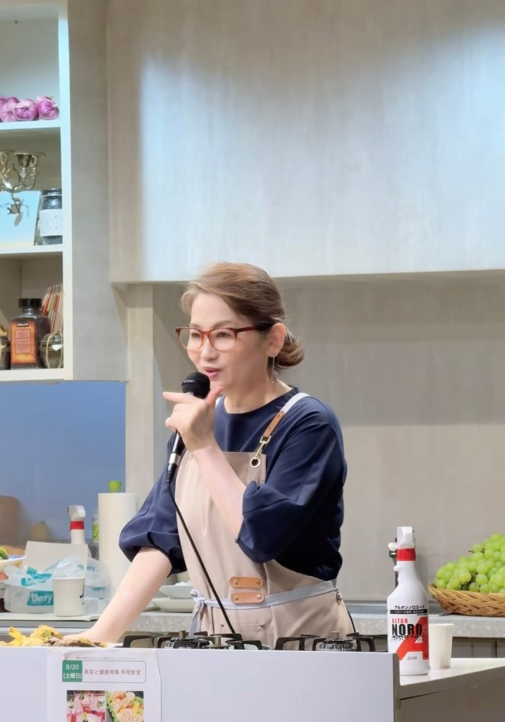 요리 연구가가 마이크를 들고 발표하는 모습, 배경에 주방 용품과 식재료가 보임.