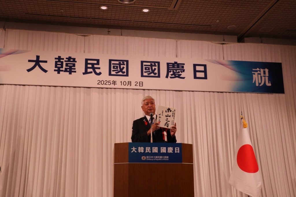 A speaker presenting at a podium during a reception celebrating the 4357th anniversary of Gaecheonjeol and the 77th Armed Forces Day at a hotel in Tokyo, with a banner in the background and flags of South Korea and Japan displayed.