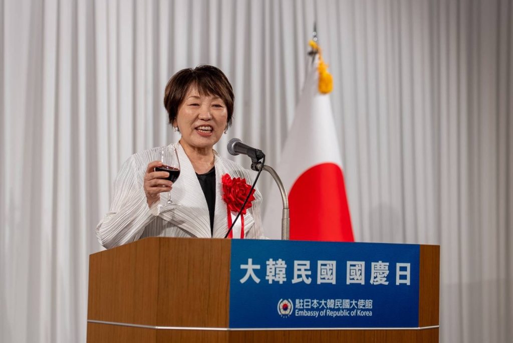 A female speaker at a podium holding a glass, smiling and delivering a speech during a national day reception at the South Korean embassy in Japan, with flags in the background.