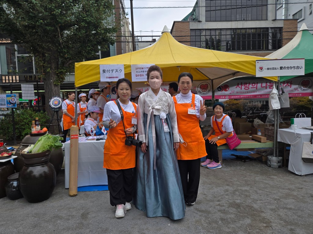 A group of women dressed in traditional Korean hanbok and bright orange volunteer shirts pose together at a festival booth, with various event activities happening in the background.