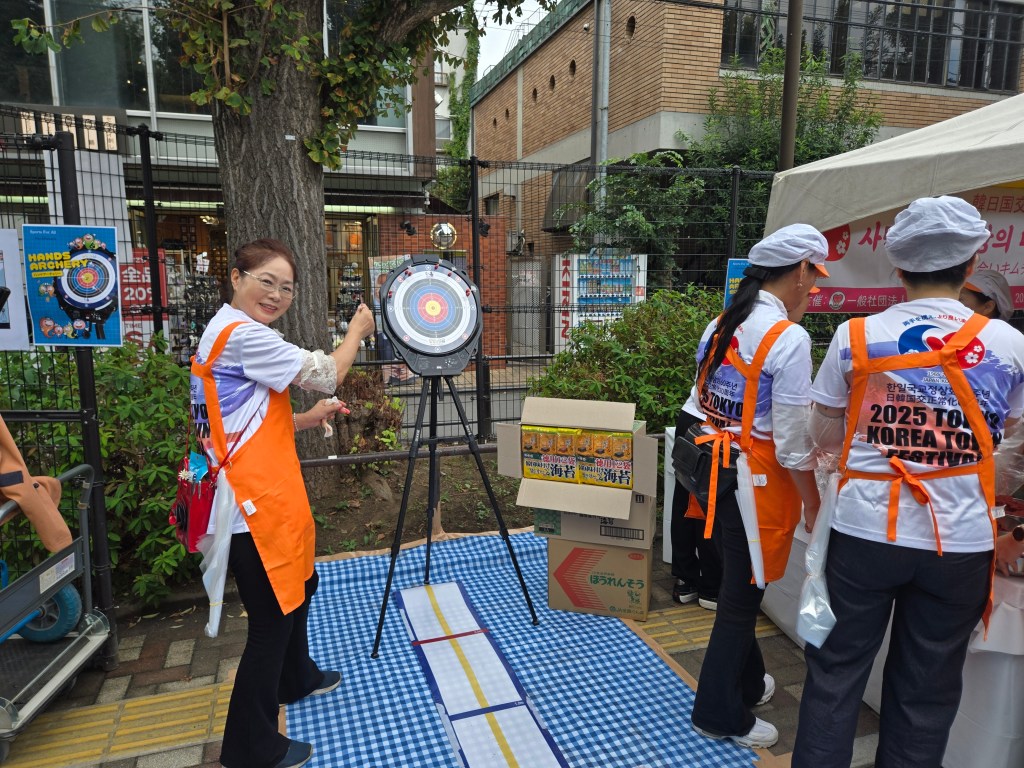 A woman in an orange apron is participating in an archery activity at the 2025 Tokyo Korea Town Festival, smiling as she prepares to throw an arrow at a target. In the background, other participants are engaged in various activities, and festival decorations are visible.