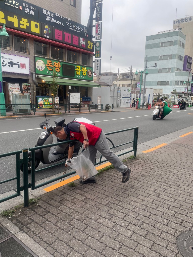 한 남성이 거리에서 쓰레기를 줍고 있는 모습. 배경에는 음식점과 상점들이 있는 신오쿠보 거리가 보인다.
