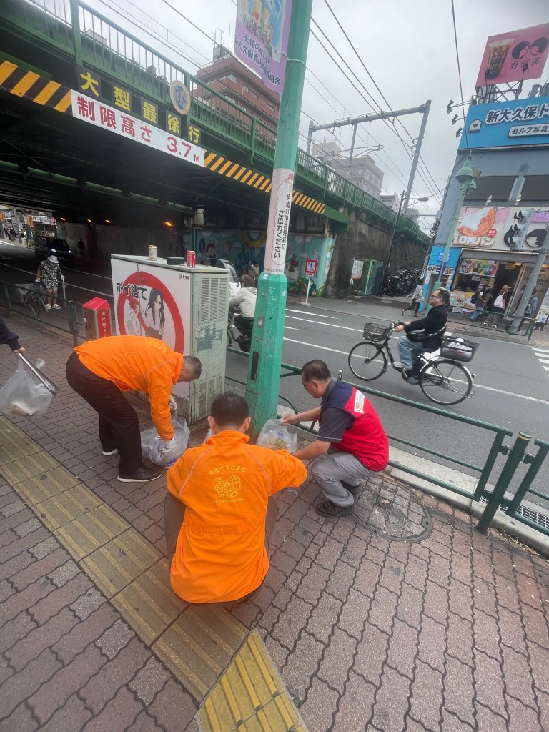 두 명의 사람들이 도로에서 쓰레기를 줍고 있는 모습, 한 명은 주황색 작업복을 입고 있으며, 다른 한 명은 빨간색 조끼를 입고 있다. 주변에는 도시 풍경이 보인다.