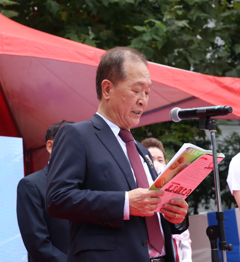 A person reading from a document while speaking into a microphone at an outdoor event, under a red tent.