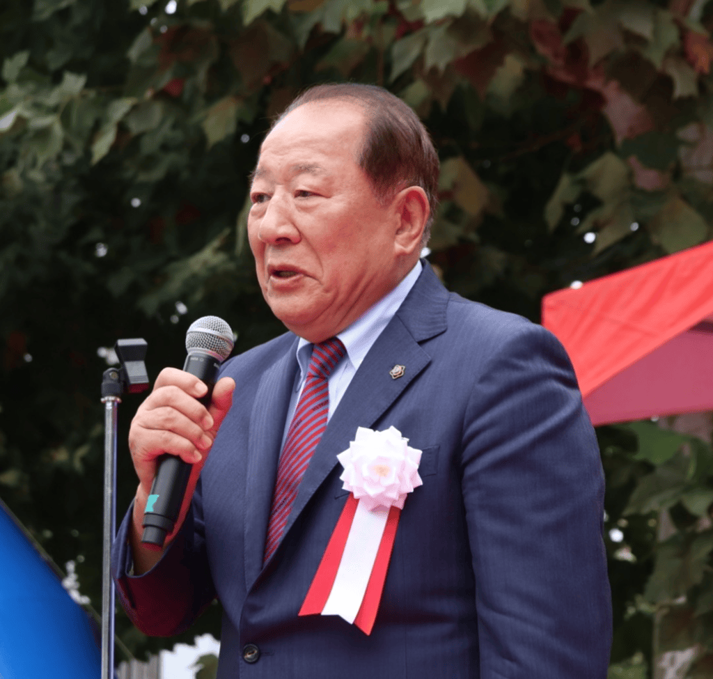 한 행사가 진행 중인 모습으로, 연단에서 마이크를 들고 있는 중년 남성이 등장하고 있다. 배경에는 나무와 장식물이 보인다.