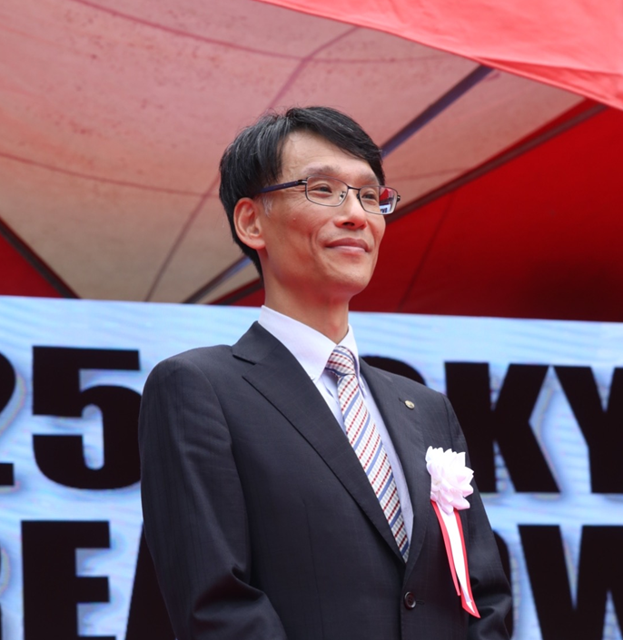 A man in formal attire standing on stage during the 2025 Tokyo Korea Town Festival, smiling with a backdrop displaying the event's name.