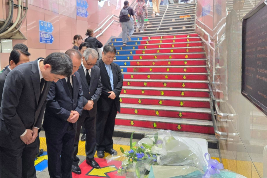 A group of people in formal attire bowing in remembrance at a memorial site in a train station, with flowers laid on the ground and colorful steps in the background.