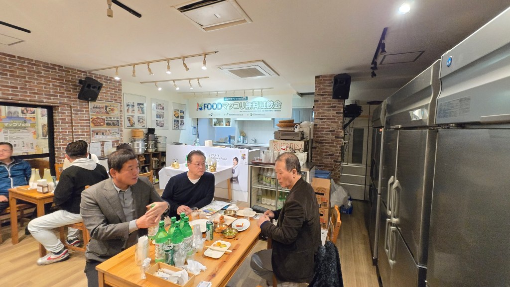 A gathering at a Korean restaurant in Shinjuku, Tokyo, during the 'K-FOOD 막걸리 무료 시음회', showcasing participants enjoying traditional Korean food and drinks in a warm atmosphere.