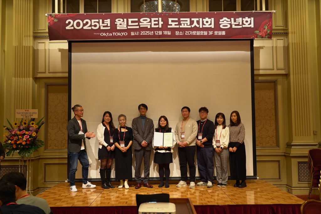 2025년 월드옥타 도쿄지회 송년회에서 참석자들이 단체 사진을 찍고 있는 모습. 배경에는 행사 배너가 걸려 있으며, 참가자들은 기념품을 들고 있습니다.