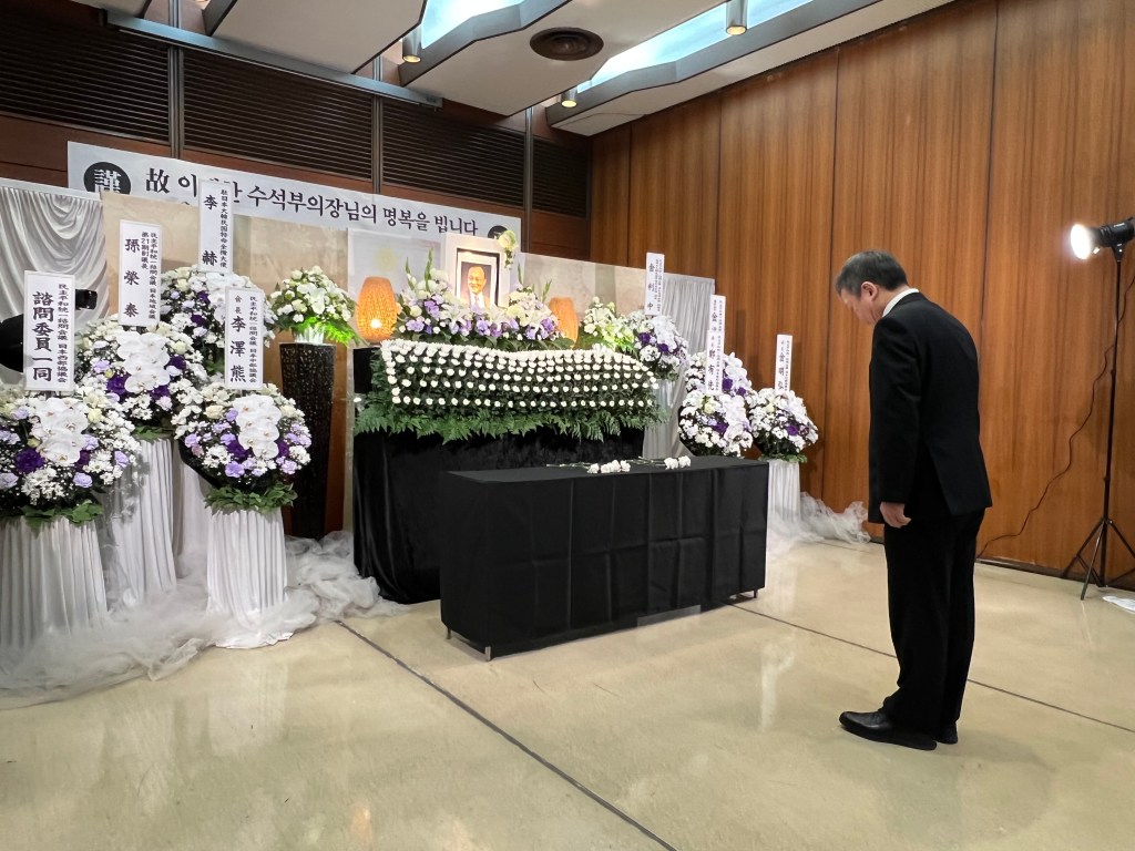 장이 없는 장례식 장면, 검은 슈트를 입은 남성이 헌화하며 고개 숙이고 있음. 잔디와 흰색 꽃으로 장식된 위패와 환영의 시민들이 보여짐.