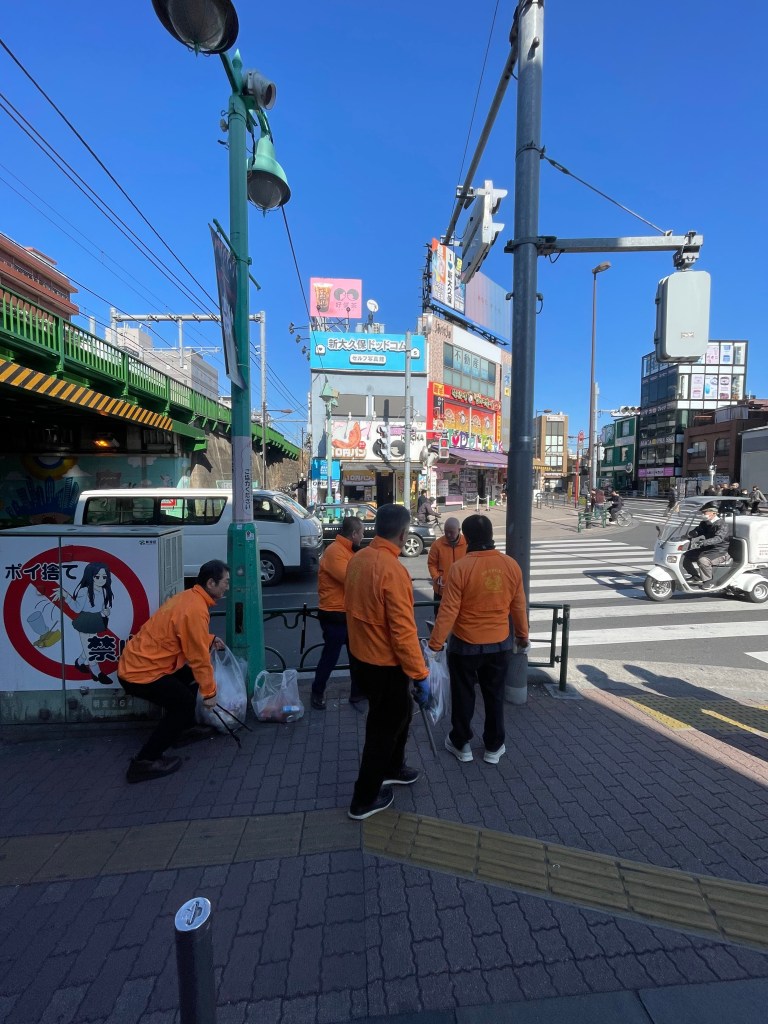 사람들이 도로변에서 쓰레기를 줍고 있는 모습. 밝은 하늘 아래, 오렌지색 작업복을 입은 여러 명이 모여 있습니다.