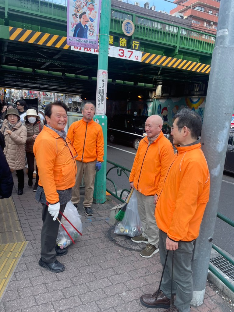 다섯 명의 사람이 주황색 점퍼를 입고 손에 쓰레기 봉투를 들고 대화하는 모습.